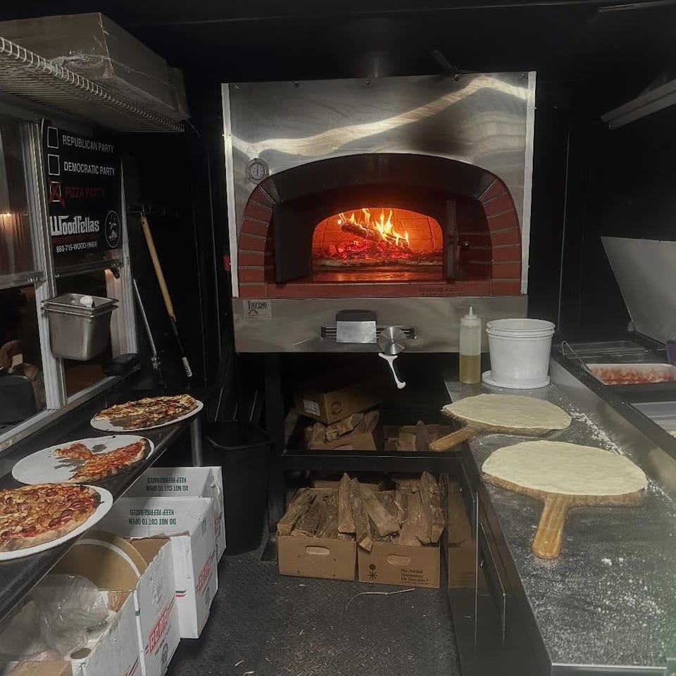 Restaurant kitchen with a wood-fired pizza oven, topped pizzas on plates, dough rounds, and ingredient trays.