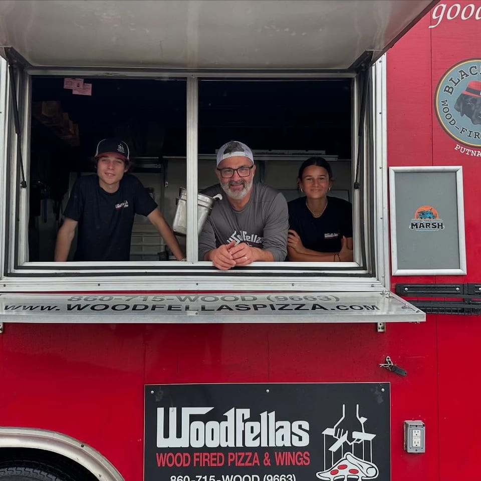 Three Woodfellas staff members pose at the service window of a red wood-fired pizza truck.