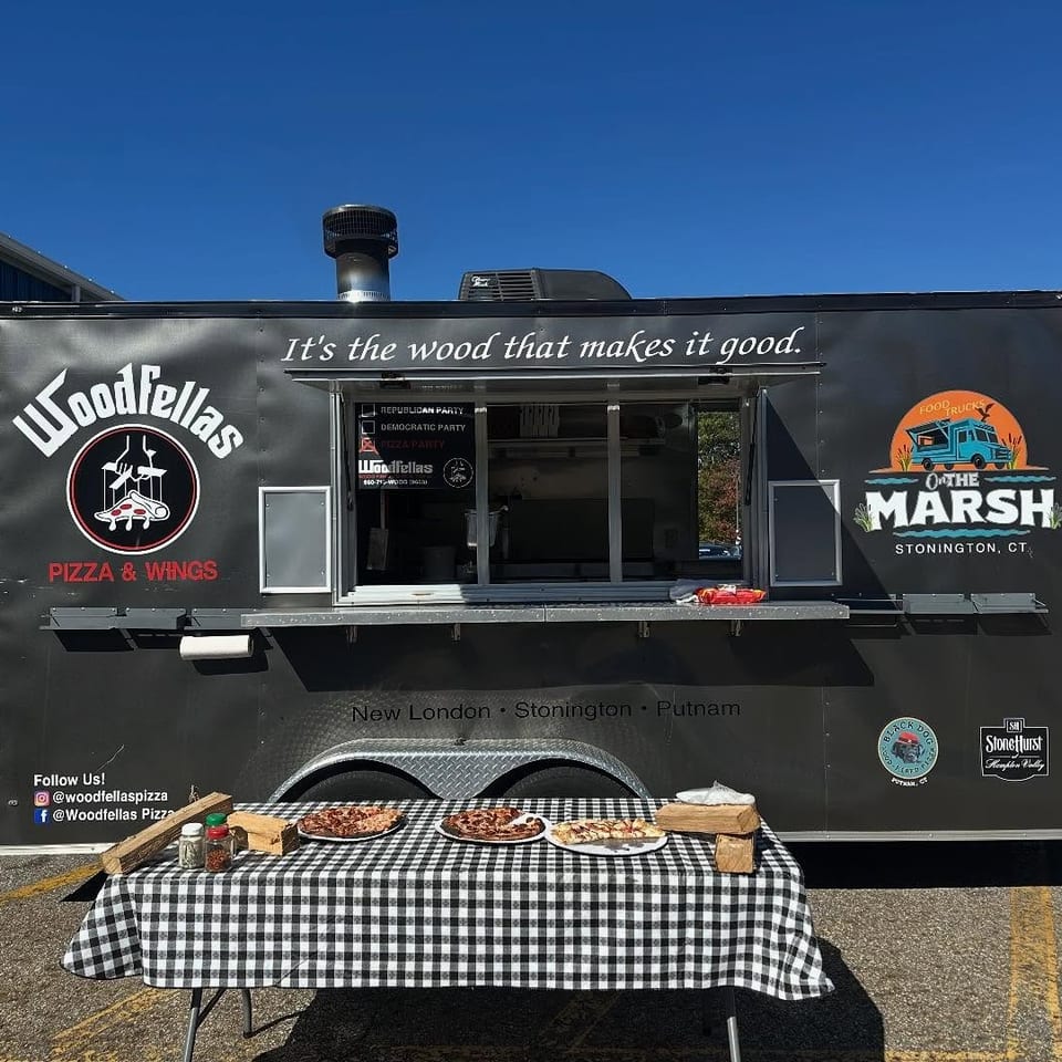 Woodfellas Pizza & Wings food truck with serving window and checkered table displaying pizzas, condiments, and wooden serving boards.