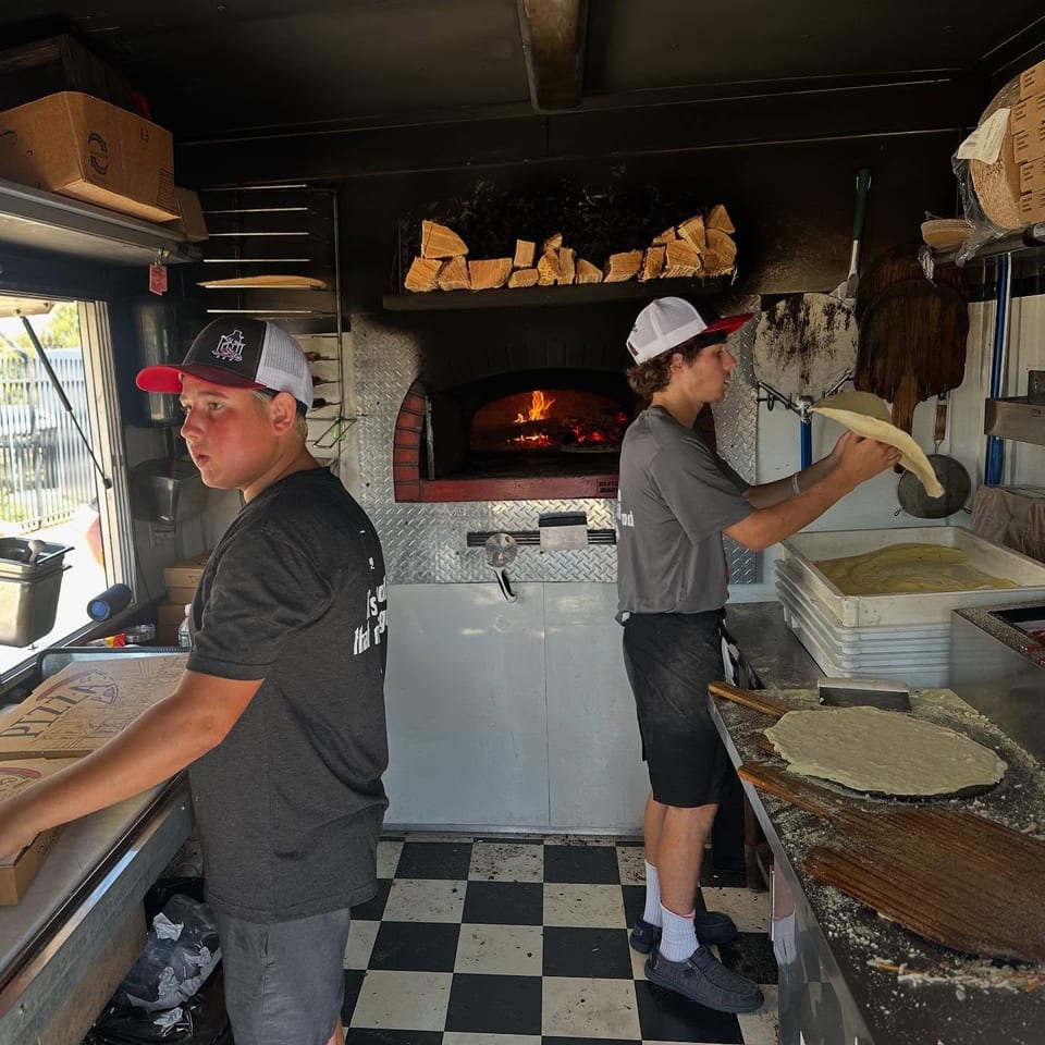 Two workers prepare flatbread inside a food trailer, with a wood-fired oven, dough, and flour-covered counter visible.