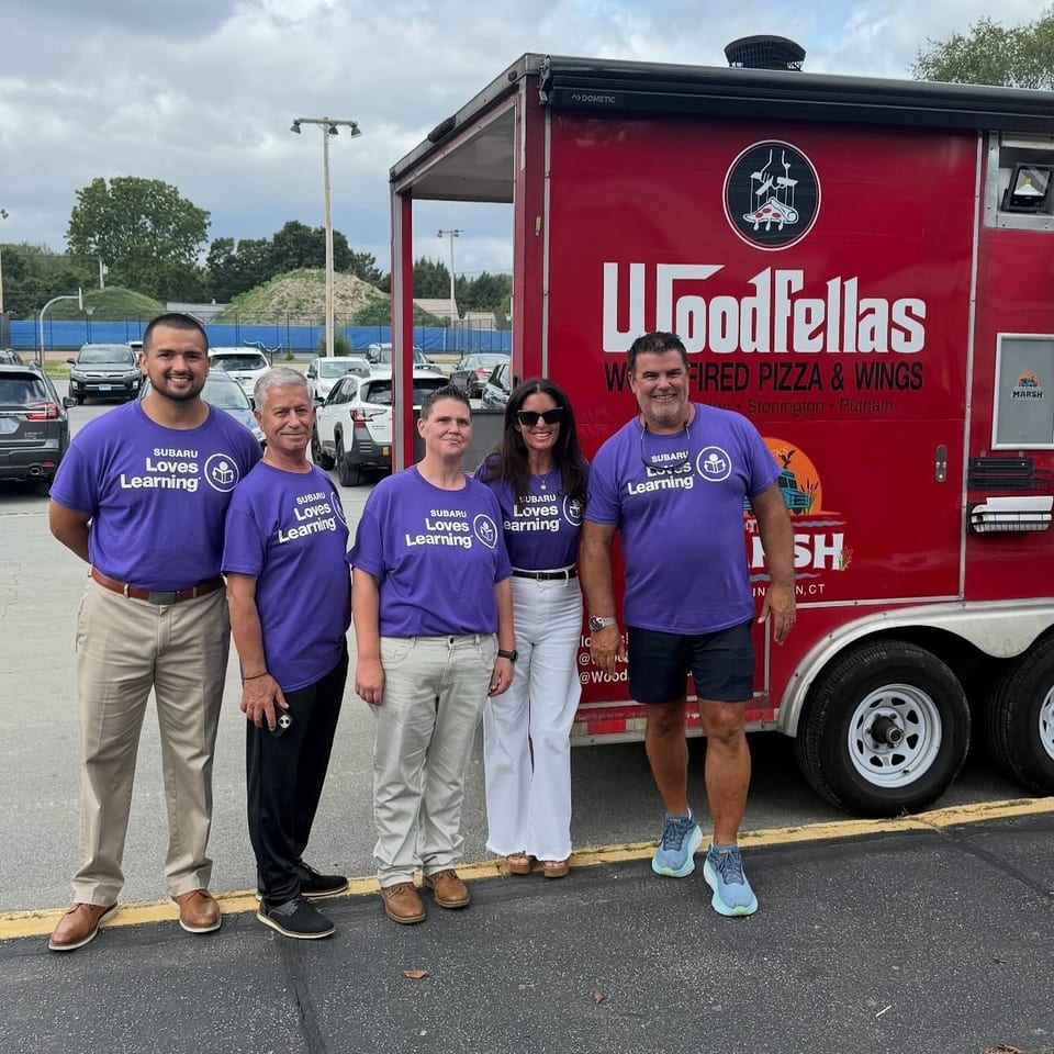 Five people wearing purple Subaru Loves Learning shirts pose beside a red Woodfellas wood-fired pizza and wings food truck.