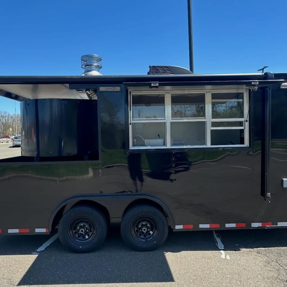 Black enclosed food trailer with serving windows, dual wheels, and open awning parked in a sunny parking lot.